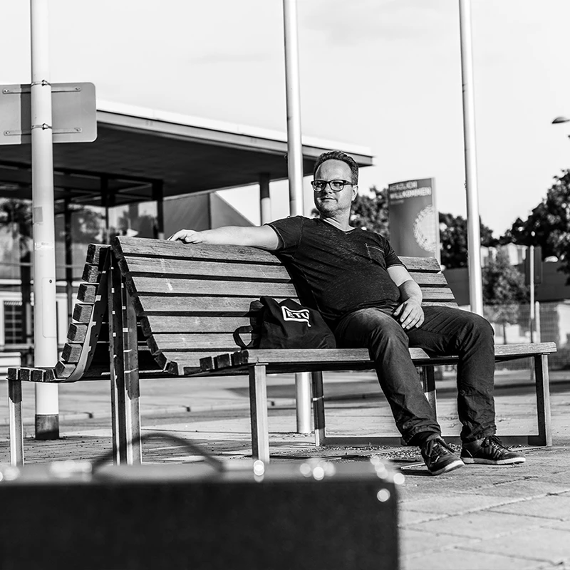 the artist Insect O. sitting on a bench with a modern glass building in the background